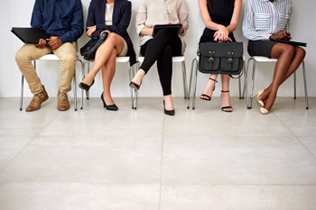Ein Mann und vier Frauen in Bürokleidung sitzen auf Stühlen nebeneinander in einer Reihe, ihre Köpfe werden vom Bildausschnitt abgeschnitten