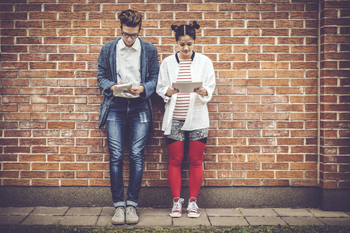 Zwei Studenten lehnen mit dem Rücken an einer Mauer. Einer notiert etwas auf einem Block, die andere blickt auf ihr Tablet.