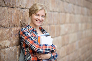Studentin lehnt mit dem Rücken an einer Mauer, in den Händen hält sie einen Stapel Bücher.