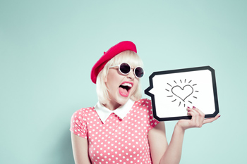 Studentin steht mit geöffnetem Mund vor einer Wand und hält eine Sprechblase in der Hand, in der ein Herz zu sehen ist.